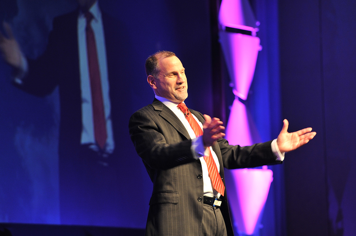 Speaker in a suit gesturing on stage, possibly discussing "Why Me?" during a conference.