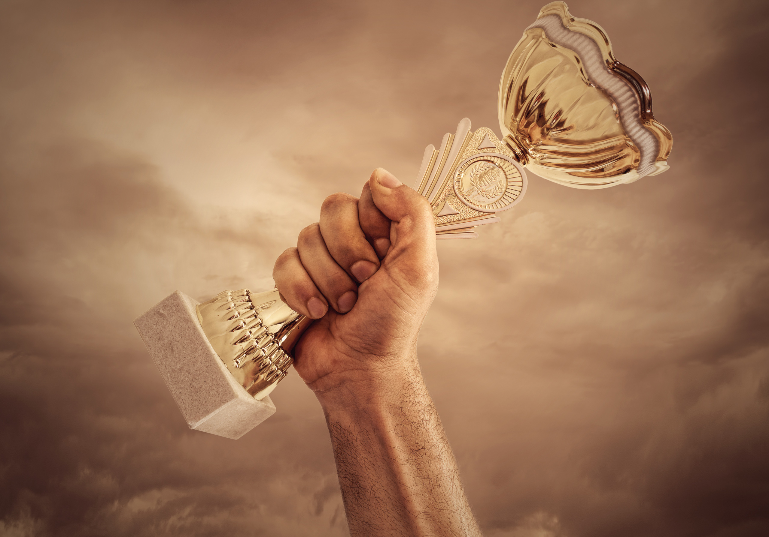 Hand holding a gold trophy against a dramatic sky. Earn the right to celebrate success.