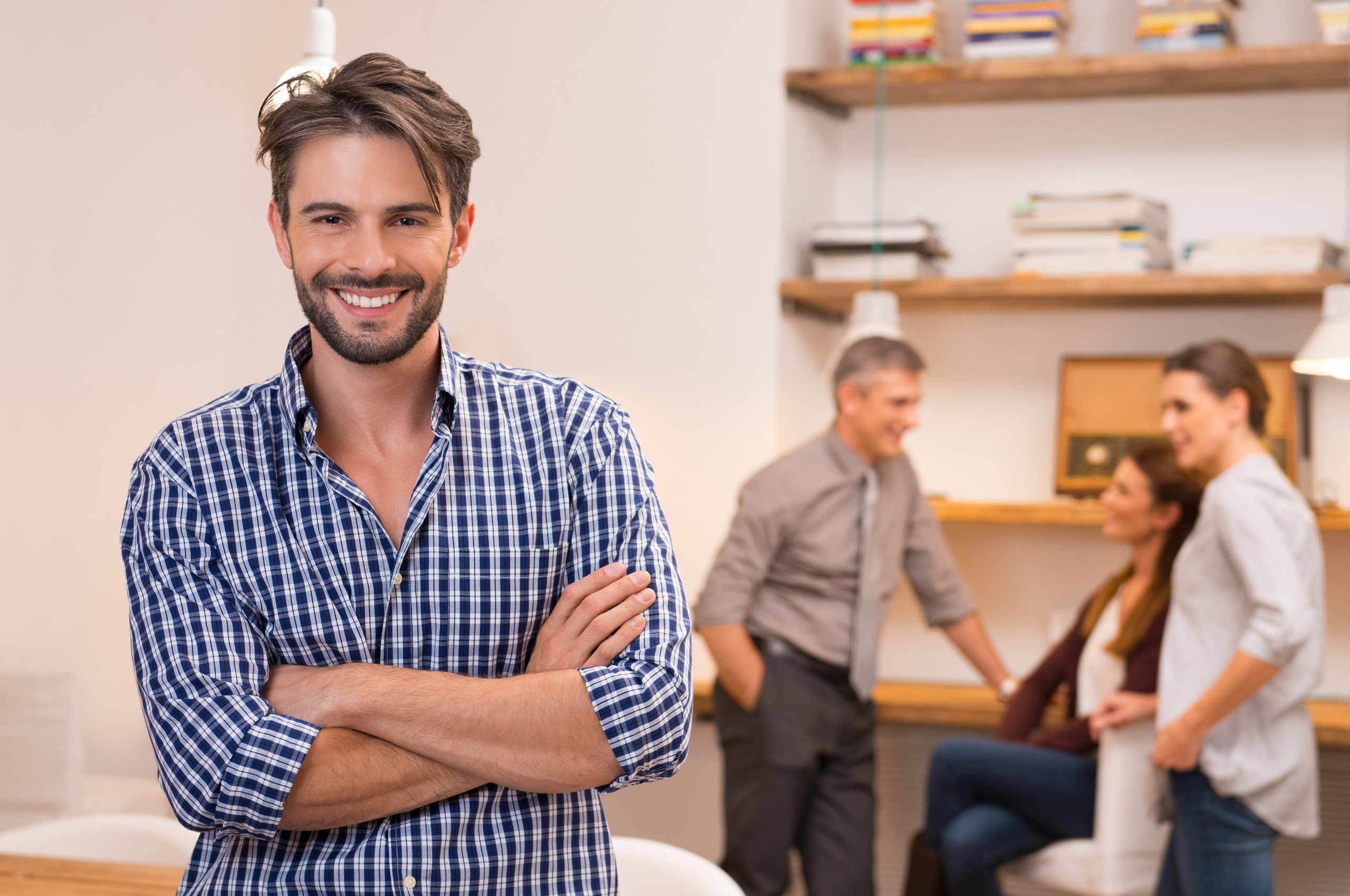 Smiling man with crossed arms in a modern office setting with colleagues.