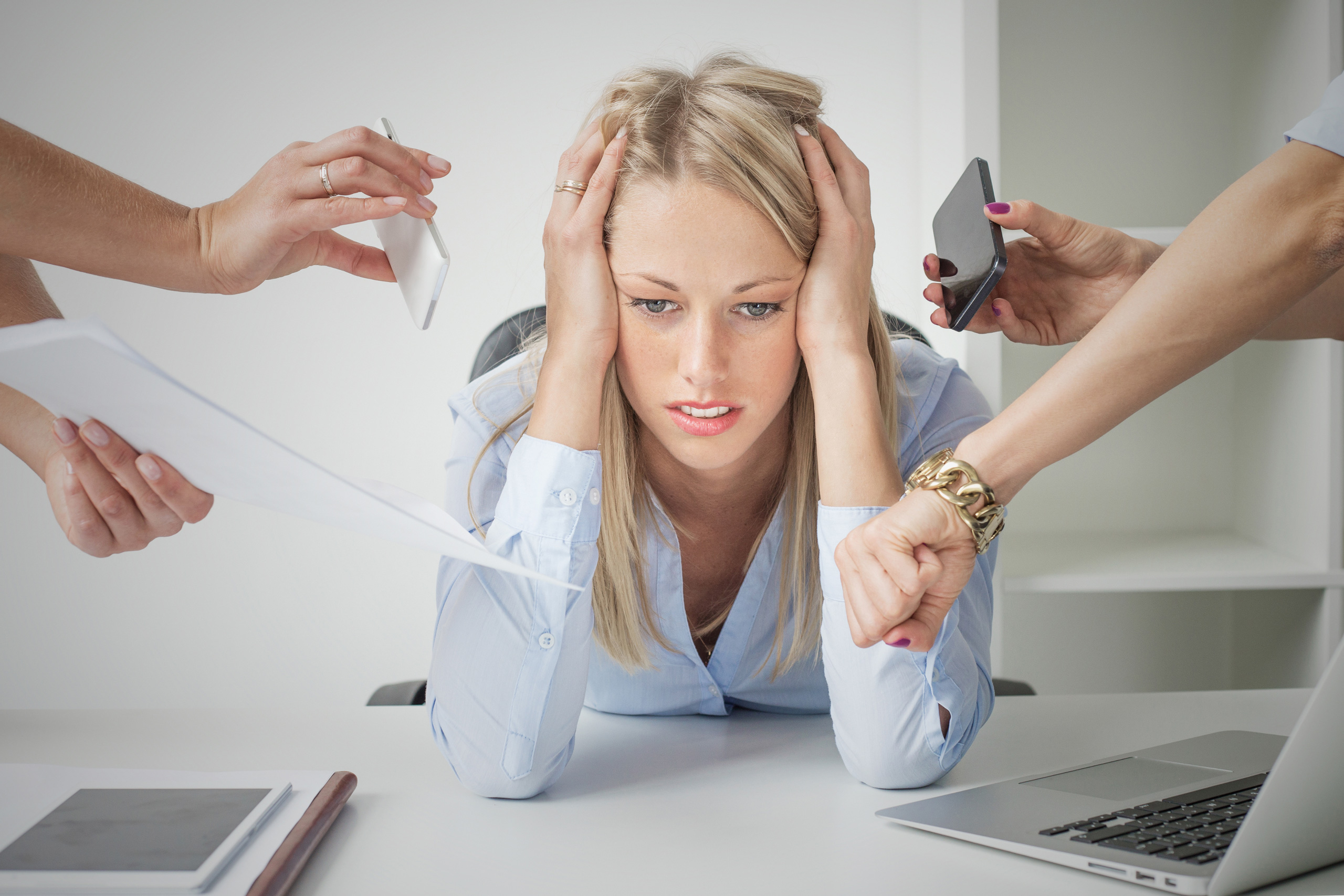 Stressed blonde woman overwhelmed at her desk with hands offering phone, documents, and reminders.
