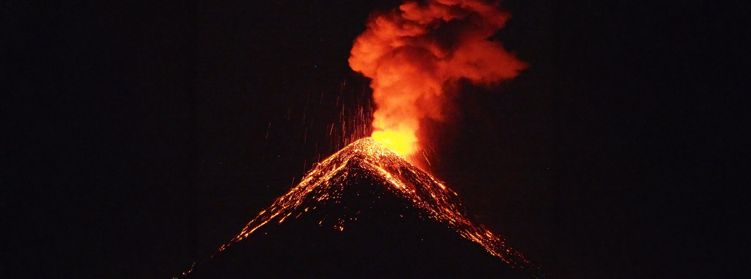 Volcano erupting at night, with flowing lava and a plume of smoke rising into the dark sky.